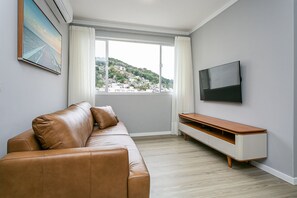 Un salon moderne avec un canapé en cuir, un téléviseur à écran plat, un meuble TV en bois et une grande fenêtre donnant sur une colline.