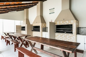 An outdoor dining area with wooden tables and benches, a brick fireplace, and a chimney. - 200m from Beira-Mar | Pool | Dining #CA107 (Florianópolis)