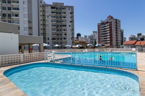 Ein Pool auf dem Dach mit Fliesenbelag, umgeben von einem Zaun und Sonnenschirmen, vor der Kulisse hoher Gebäude.
