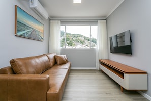 A modern living room with a leather sofa, a flat-screen TV, a painting, and a view of a hillside. - 200m from Beira-Mar | Pool | Dining #CA107 (Florianópolis)