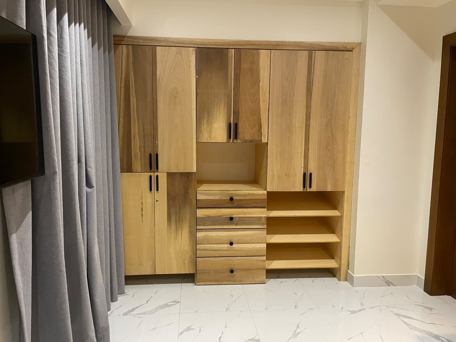 Una habitación con armario de madera empotrado, televisor de pantalla plana montado en la pared y piso con baldosas de mármol.