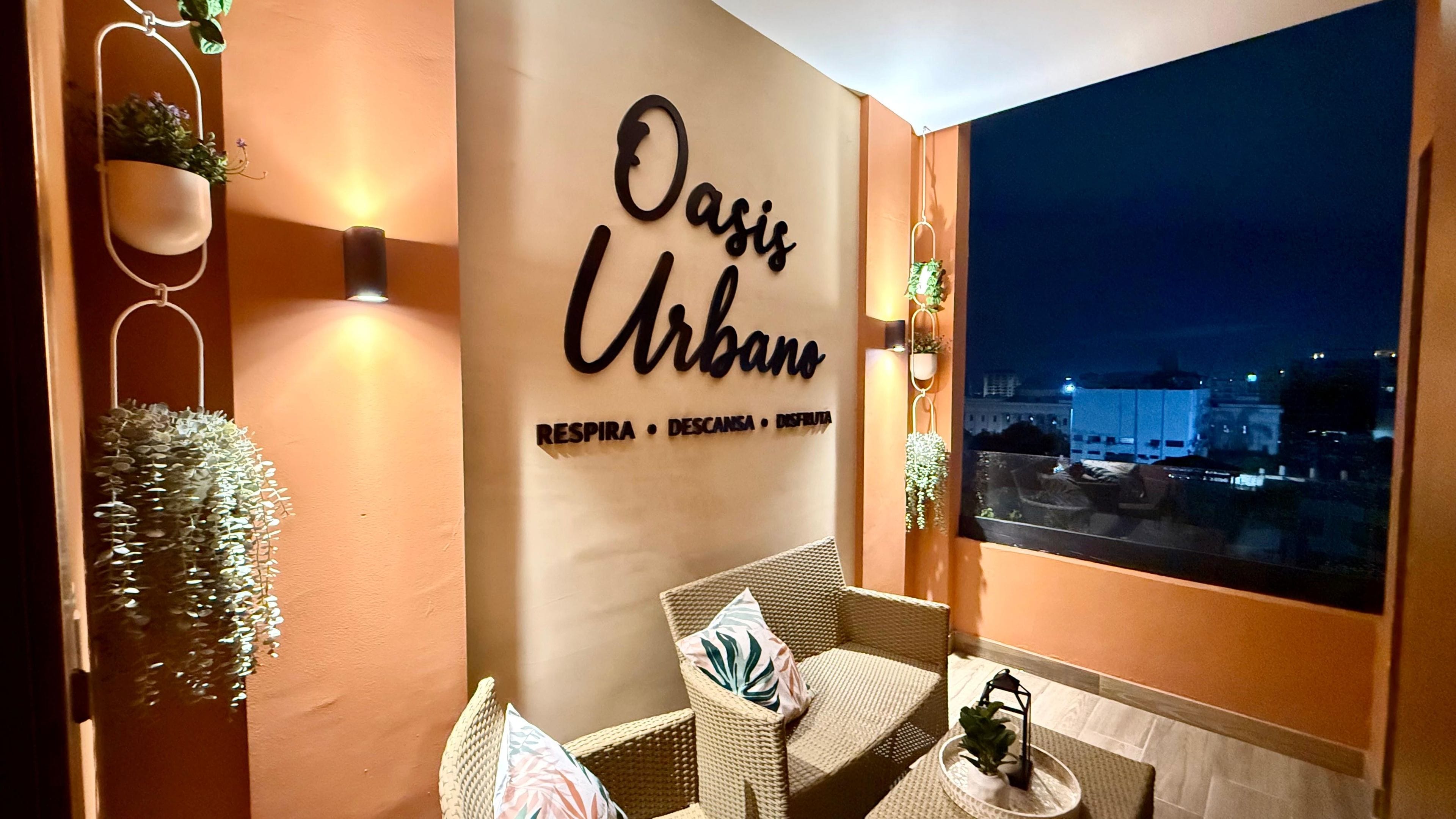 A cozy seating area with wicker furniture and a wall-mounted sign reading 'Oasis Urbano' with amenities listed.