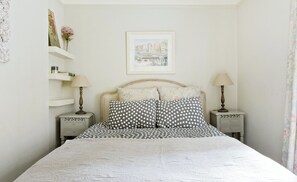 Une chambre à coucher avec un lit, deux tables de chevet, une commode, une lampe, un tableau encadré au mur et un rideau à motifs floraux.