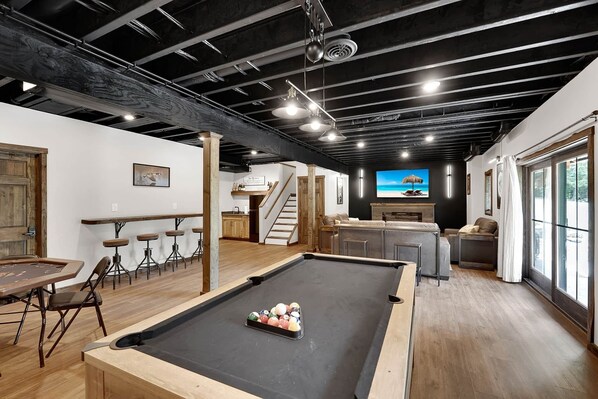 Une table de billard dans une pièce qui comprend un bar et un téléviseur diffusant des images d’une plage.
