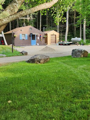 Eine Holzhütte mit Kiesauffahrt, einer blauen Tür und einem kleinen Geräteschuppen in bewaldeter Landschaft.