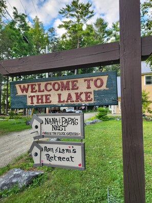 Ein hölzernes Wegweiser mit mehreren Schildern, darunter 'WILKOMMEN AM SEE', 'DAS NEST VON OMA & OPA' und 'Ron & Lori's Retreat'.