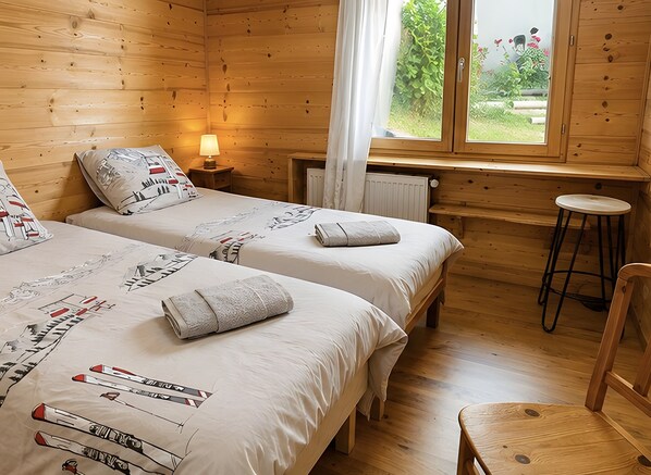 Habitación tipo cabaña de madera con dos camas, una ventana con cortinas, un radiador y una silla de madera.
