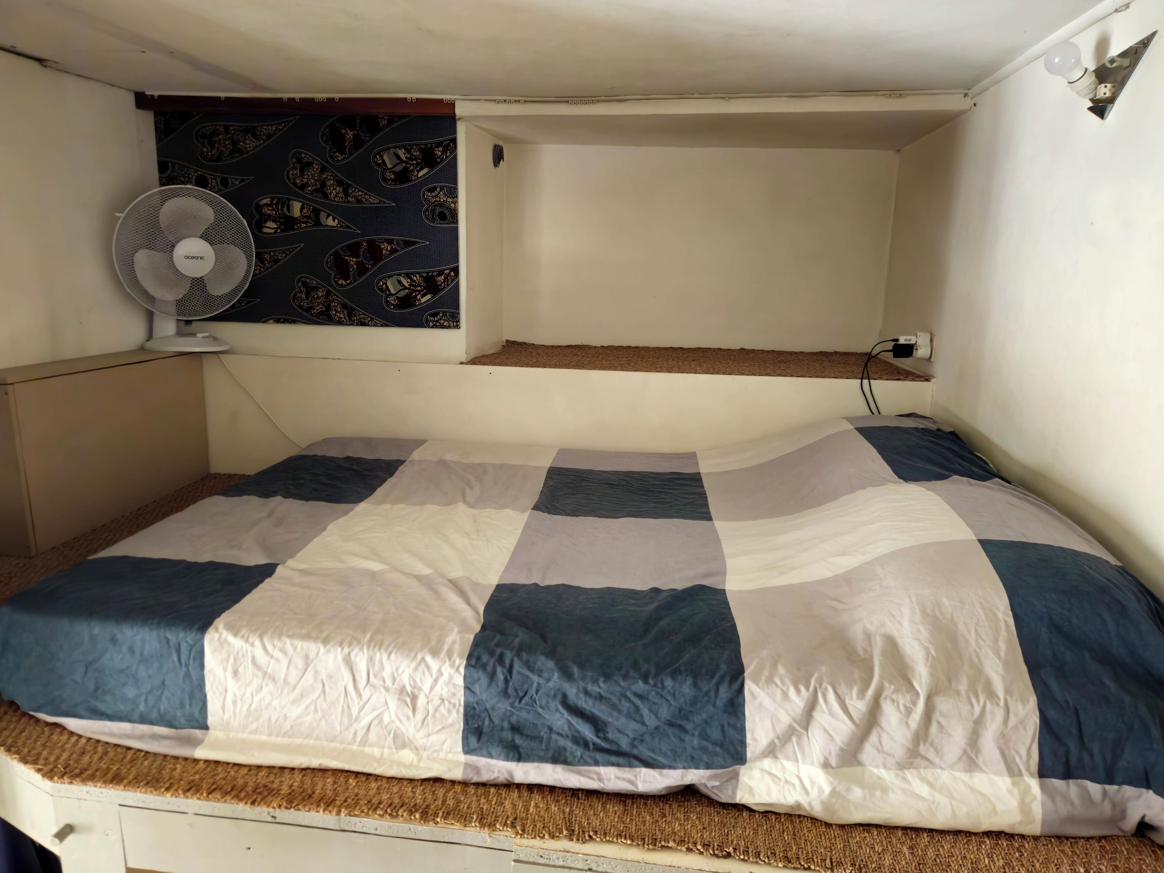 Una cama con colcha a cuadros, un ventilador de pared y un tablón de corcho.