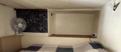 A bed with a checkered bedspread, a wall-mounted fan, and a corkboard.