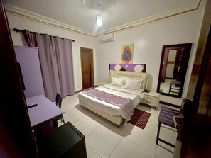 Un dormitorio con cama, mesitas de noche, un escritorio, una silla, un espejo y una puerta.