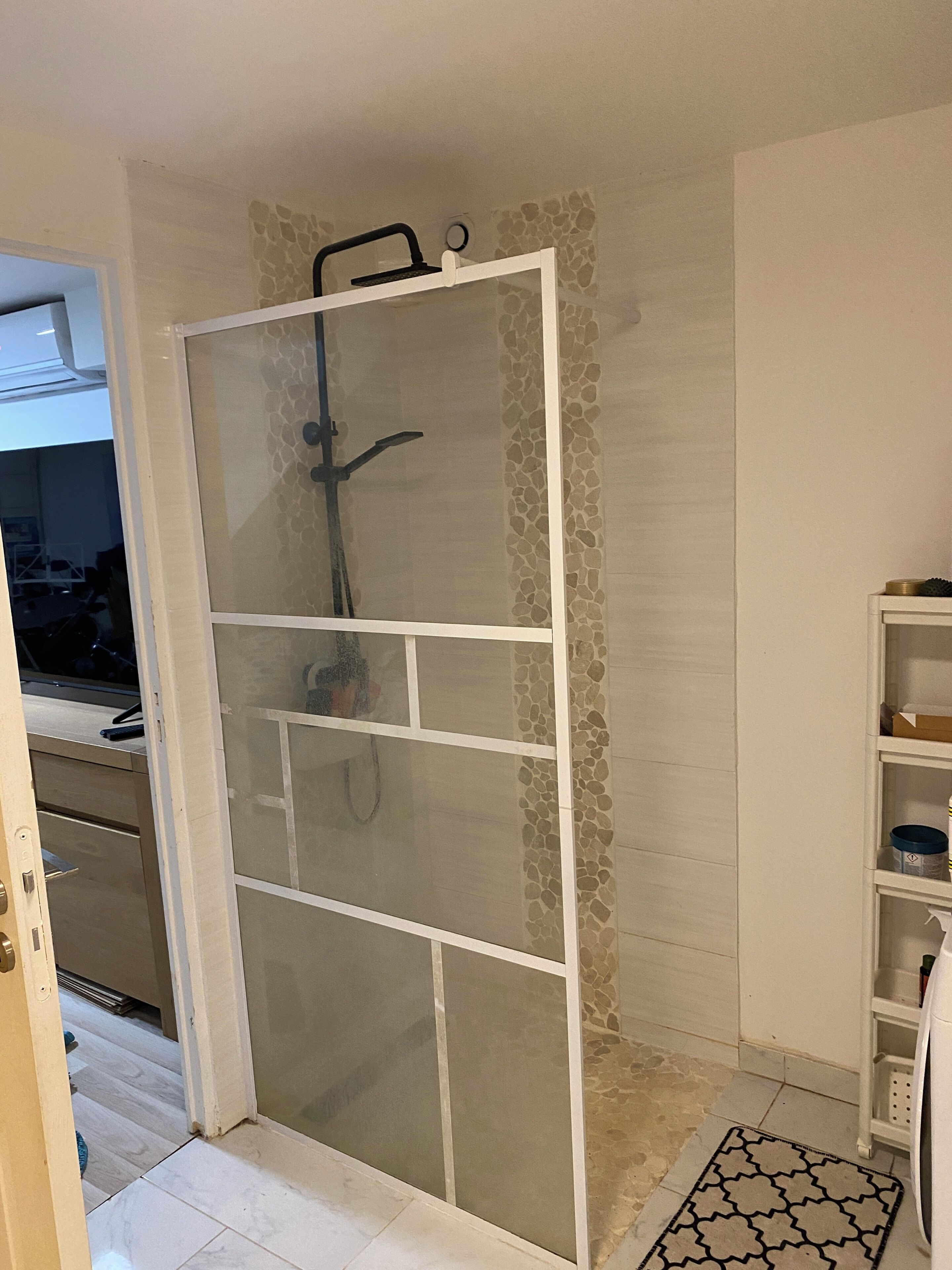 A bathroom with a shower enclosure, a tiled floor, and a white shelf unit.