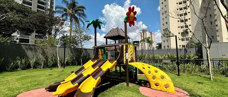 A playground with slides and climbing structures, surrounded by a fenced grassy area, with tall buildings in the background.