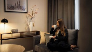 Une personne est assise sur un canapé dans une pièce. On aperçoit un tableau encadré au mur, un vase avec des branches et une lampe posée sur une table d’appoint.