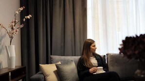 Une personne est assise sur un canapé gris, tenant une tasse blanche, dans une pièce où se trouvent un rideau foncé et un vase rempli de fleurs, posé sur une table d’appoint.