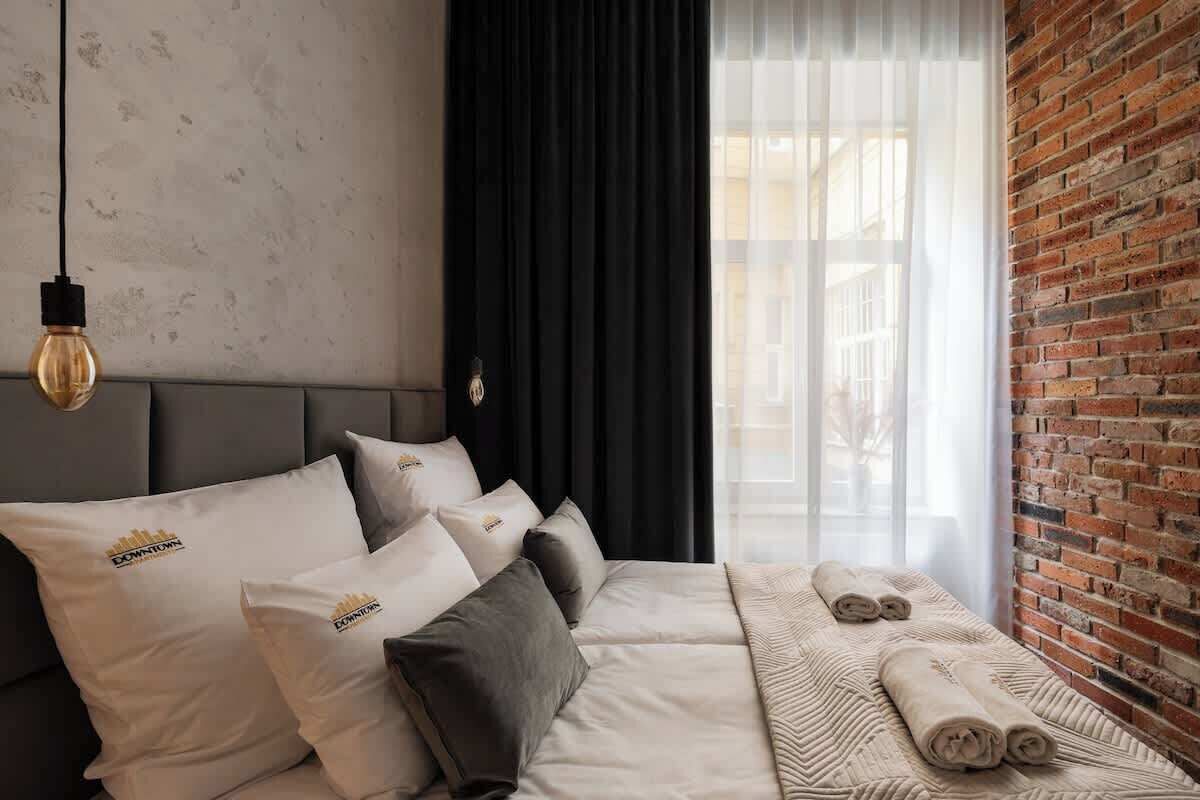 A hotel room with a bed, pillows, a blanket, a towel, a hanging light fixture, and a brick wall.