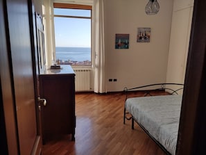A bedroom with a wooden floor, a bed with a patterned bedspread, a dresser, a radiator, and a window with a view of the sea. - Mare in Vista Salerno - Amalfi Coast Rooftop (Salerno)
