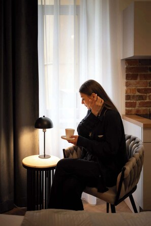 Eine Person sitzt in einem modernen Sessel an einem Tisch mit einer Lampe und hält eine Tasse, in einem Raum mit Backsteinwand und einem Fenster mit durchsichtigen Vorhängen.