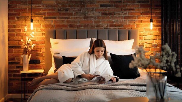 Una persona sentada en una cama leyendo un libro, con un muro de ladrillo al fondo.