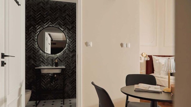 Quarto compacto com parede em padrão herringbone, um espelho redondo, uma pia e um espaço de jantar com duas cadeiras e uma mesa.