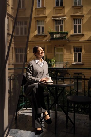 Eine Person sitzt an einem Tisch im Freien, mit einer Tasse in der Hand, und blickt auf ein Gebäude mit vielen Fenstern.