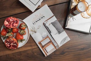 Eine Zeitschrift mit Stadtansichten auf dem Cover, eine Schale Erdbeeren und Granatapfel sowie eine Kerze auf einem Holztisch.