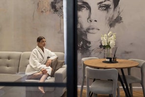 A person sitting on a sofa in a room with a large wall mural, a round wooden table with chairs, and a vase with flowers. - Charm & Chic Suite 26.2 | Main Old Town & Planty (Kraków)