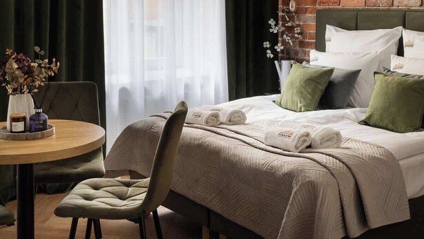 Ein Hotelzimmer mit Backsteinwand, einem Bett mit grün-weißen Kissen, einem runden Tisch mit Stühlen und einem Fenster mit transparenten Vorhängen.