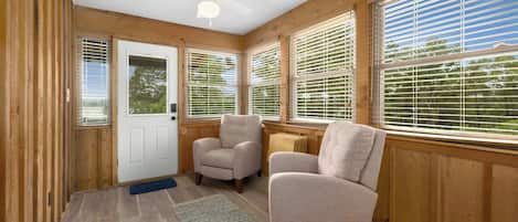 A room with wooden walls, a ceiling fan, two armchairs, a door, and large windows with blinds.