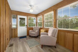 A room with wooden walls, a ceiling fan, two armchairs, a door, and large windows with blinds.