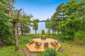 A wooden deck with four Adirondack chairs and a central fire pit, surrounded by a forest and overlooking a lake.
