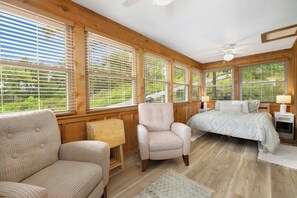 A bedroom with a bed, two chairs, a small table, and large windows with blinds.