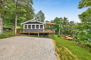A house with a deck, surrounded by trees and a gravel driveway.