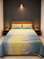Una cama con cubrecama floral, cabecera de madera, mesitas de noche con plantas y una lámpara fijada a la pared.