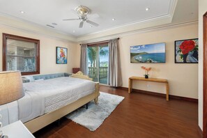 Un dormitorio con cama, ventilador de techo, ventana con persianas, un cuadro, un jarrón con flores y un banco de madera.
