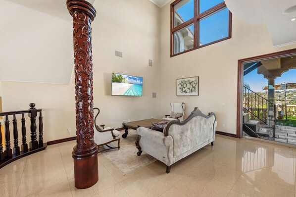 Un amplio salón con techo alto, un ventanal grande, un pilar de madera, un sofá blanco, una mesa de centro y un televisor.