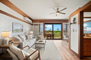 Un salon spacieux comprenant un canapé, une table basse et des fauteuils, un ventilateur de plafond et une vue sur un balcon aménagé avec du mobilier d’extérieur, accessible par une porte-fenêtre coulissante.