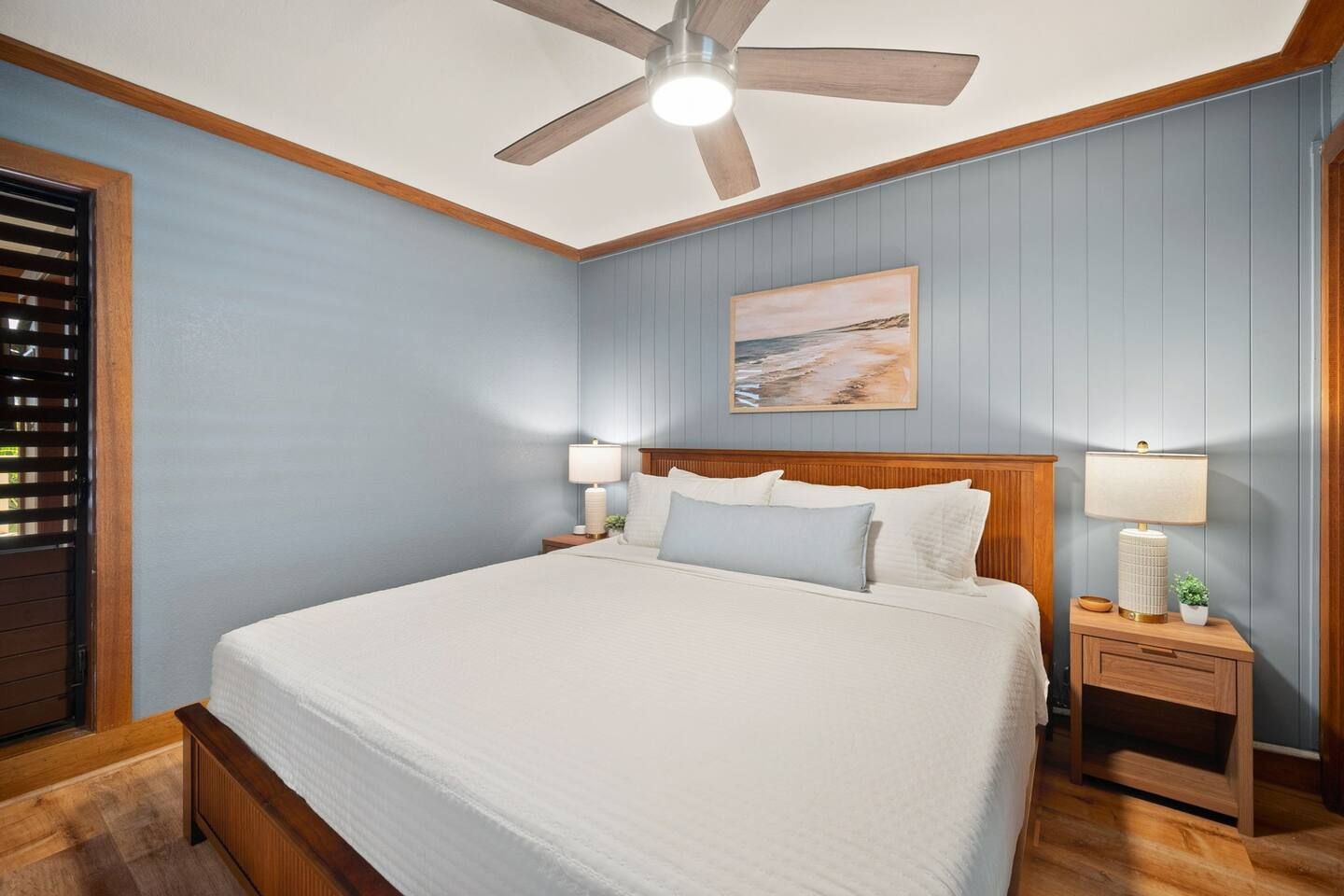 A bedroom with a bed, wooden headboard, two bedside tables with lamps, a ceiling fan, and a painting on the wall.