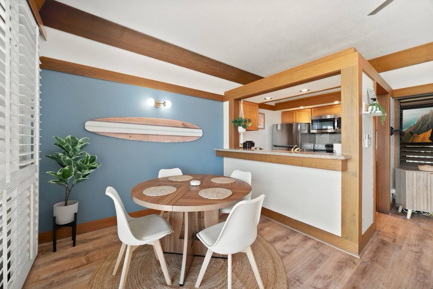 A compact kitchen with modern appliances, a dining area with a round table and white chairs, and a surfboard decoration on the wall.