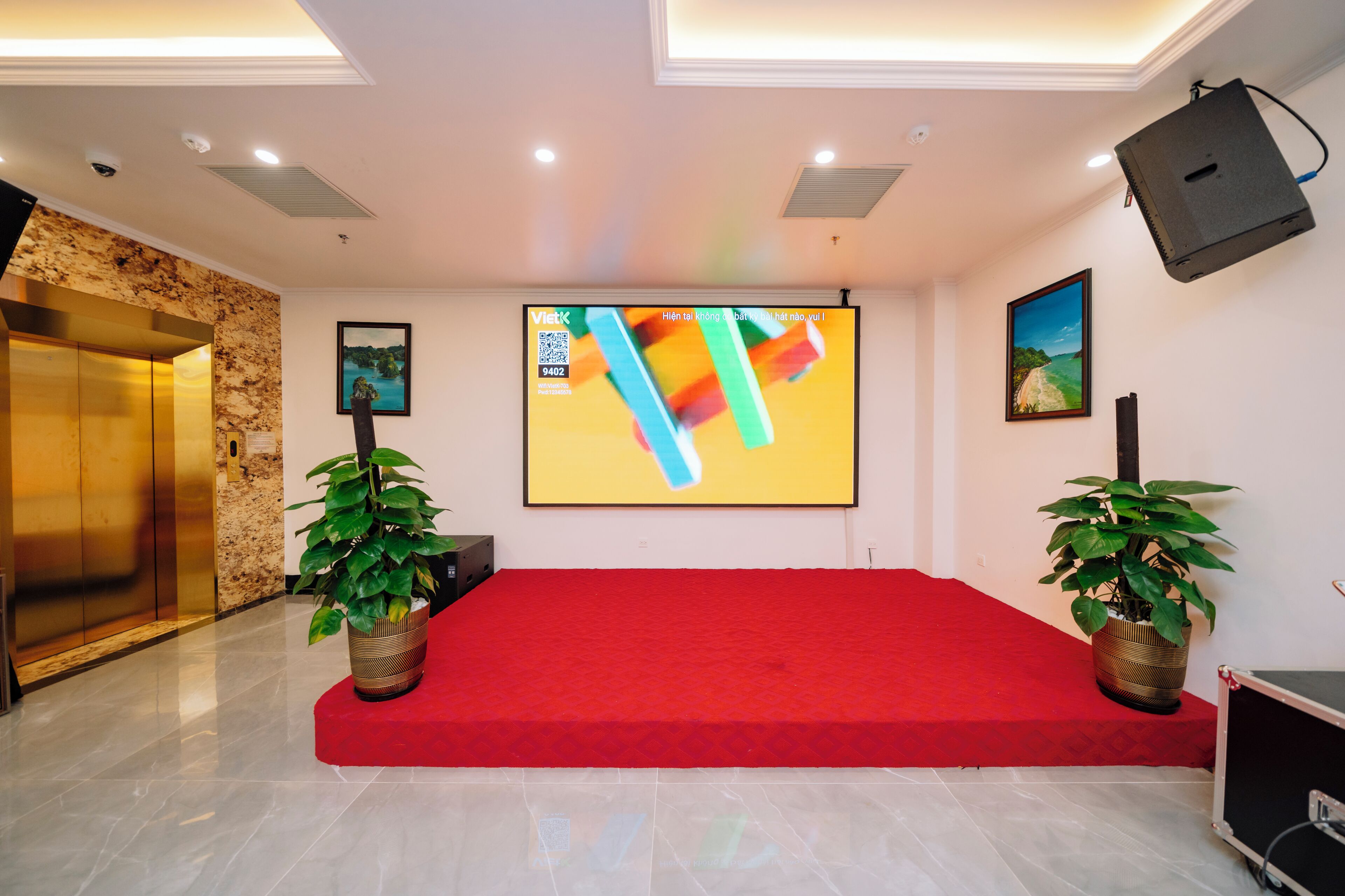 Un salón de conferencias con alfombra roja, una pantalla grande, dos plantas en macetas y un proyector.