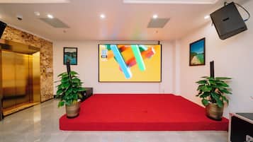 Un salón de conferencias con alfombra roja, una pantalla grande, dos plantas en macetas y un proyector.
