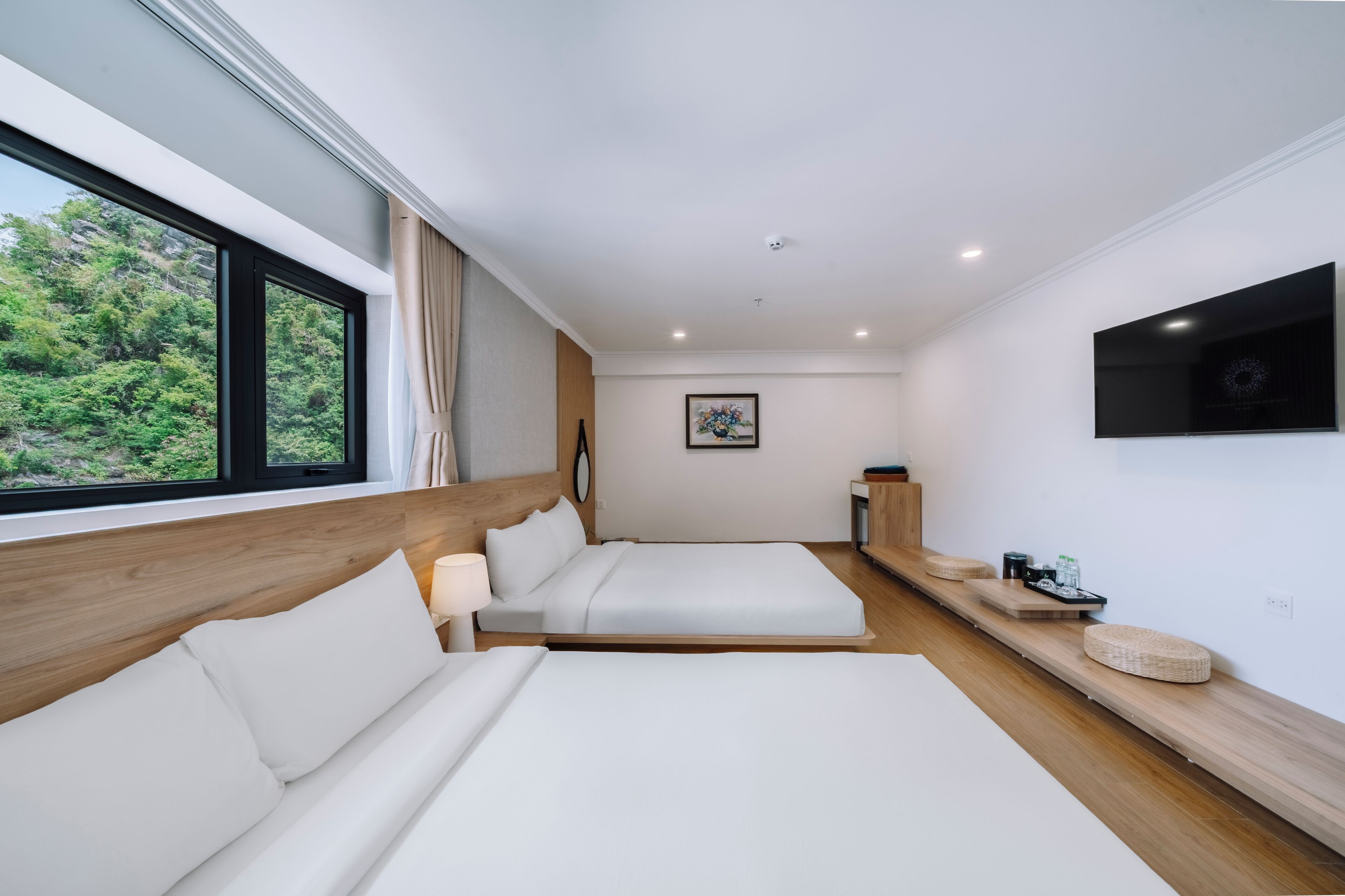 A modern hotel room with two beds, a TV, and a view of greenery outside.
