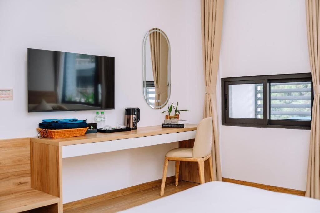 A hotel room with a bed, a desk with a television, a chair, a window with curtains, and a small table with a plant and a basket.