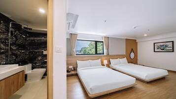 A modern hotel room with a wooden floor, a large bed, a separate bed, a bathroom with a marble wall, and a view of greenery outside.