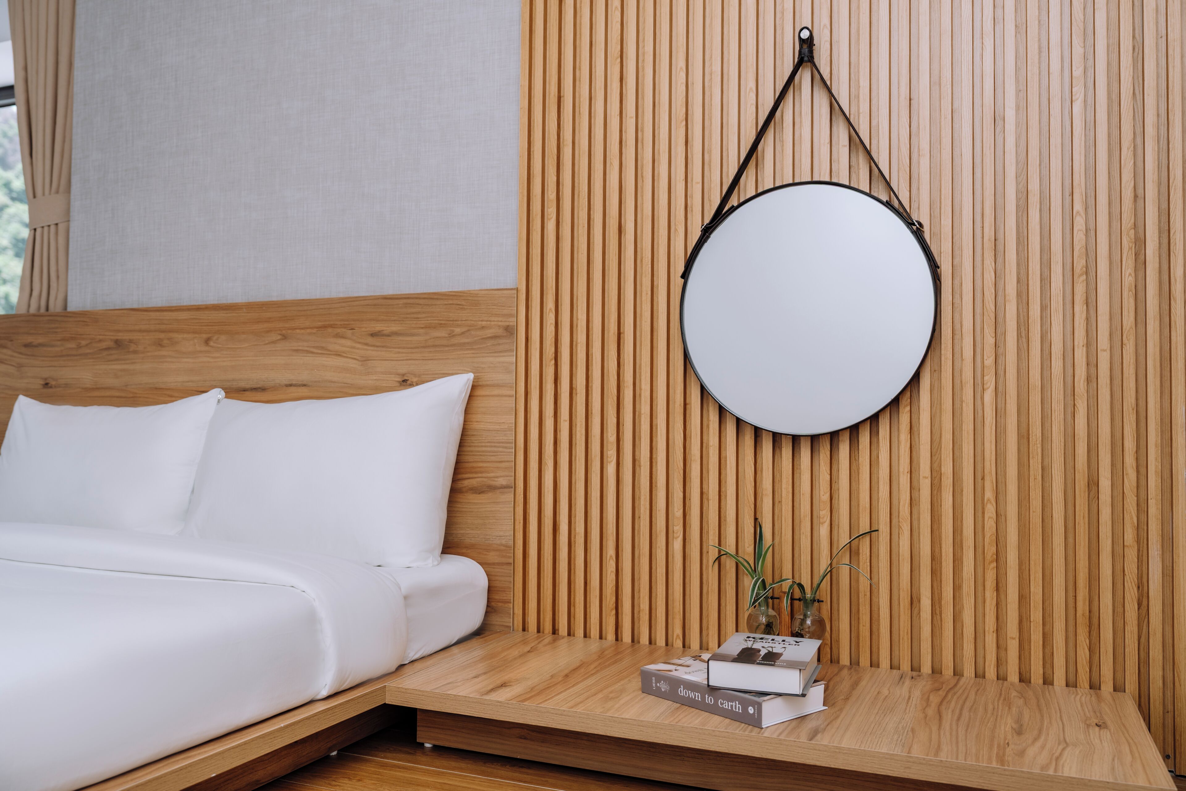 Una cama con ropa de cama blanca, un espejo redondo en la pared y una mesita con libros y una planta.