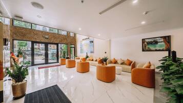 Um lobby de hotel com piso de mármore, área de estar com cadeiras laranja, um mapa grande na parede e vista para o exterior através de amplas janelas.