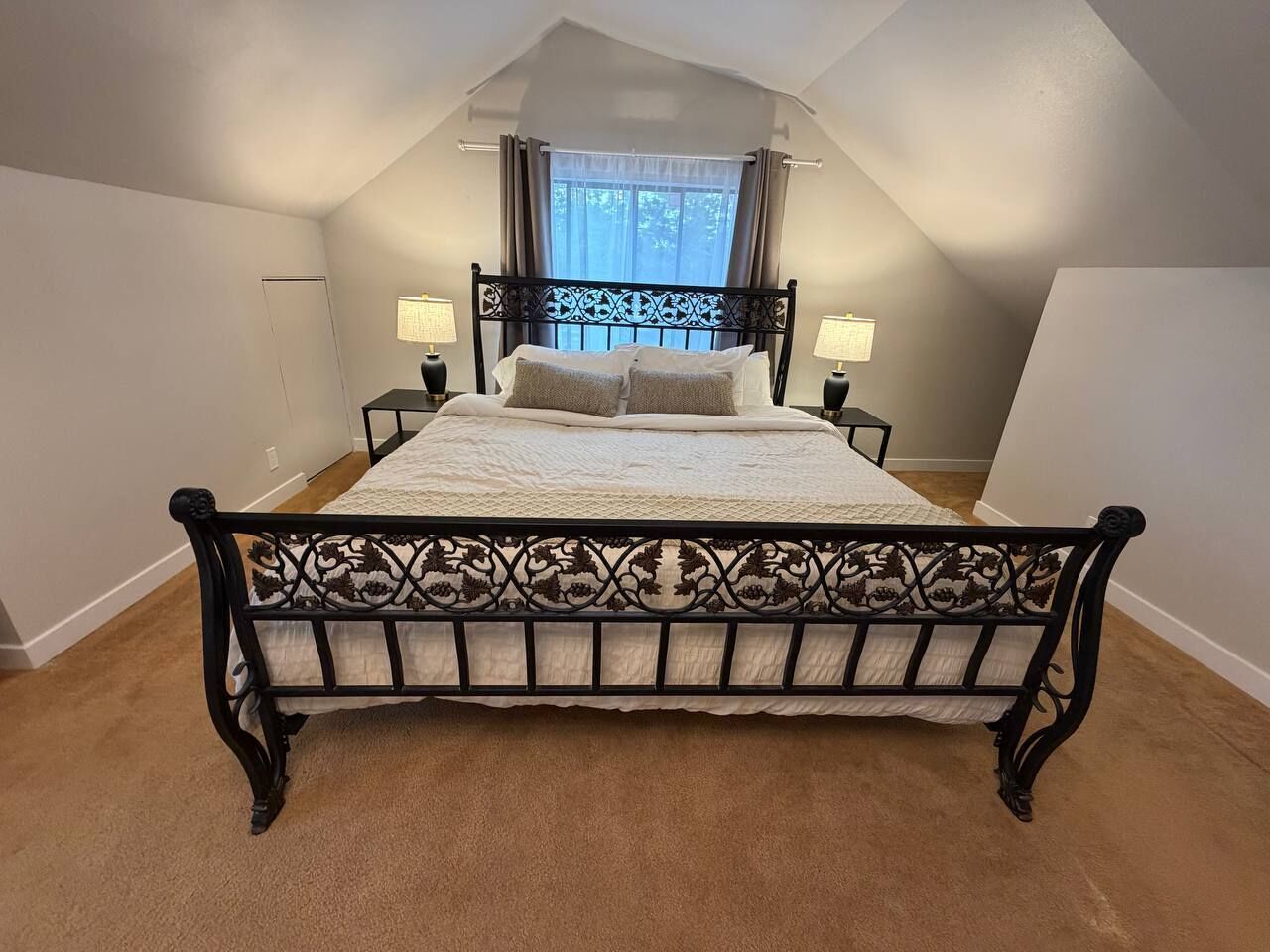A bedroom with a bed, two bedside lamps, a window with curtains, and a sloped ceiling.