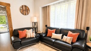 A modern living room with a black leather sectional sofa, a round wall clock, and a large window with curtains. - Mountain Cabin With Hot Tub & Fire Pit, Wallace Falls & Stevens Pass (Gold Bar)