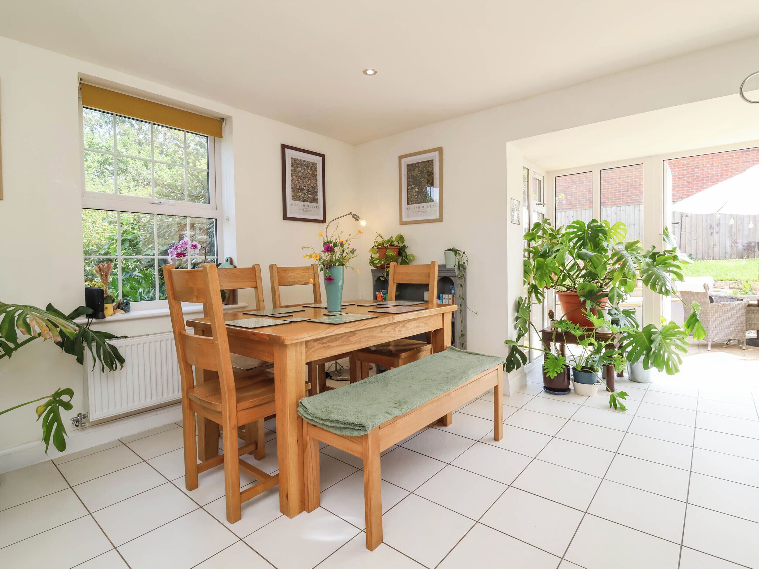 A dining area with a wooden table and chairs, a bench, potted plants, and framed artwork on the walls.
