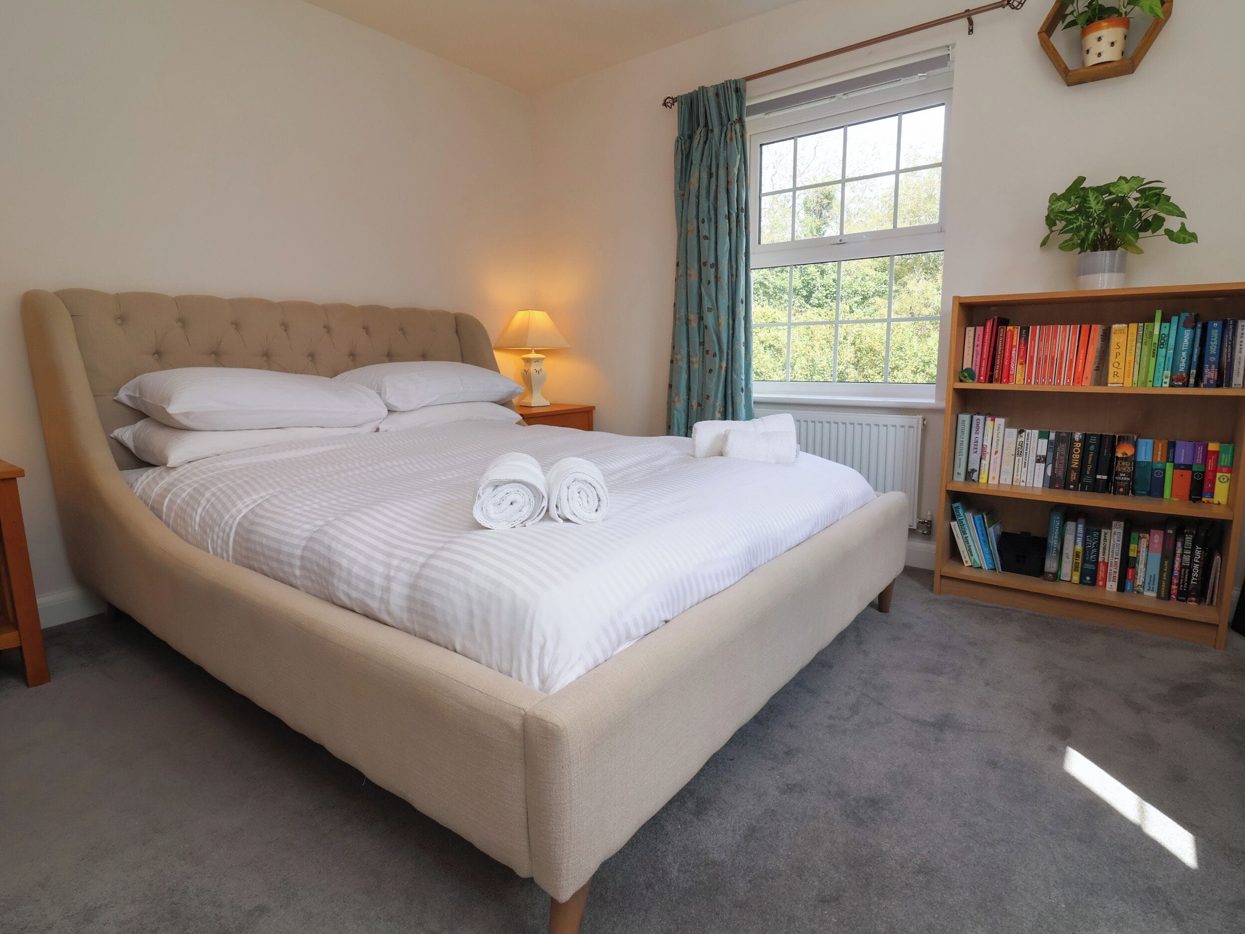 Una cama bien hecha con almohadas y toallas enrolladas, una lámpara de noche, una estantería con libros, una ventana con cortinas y un radiador.