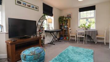 A living room with a flat-screen TV, a wooden entertainment unit, a blue ottoman, a teal rug, a bookshelf, and a dining area with a table and chairs.